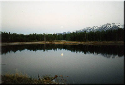 The moon sets, reflected in still water.